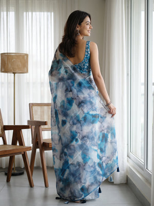 A woman wearing a blue and white watercolor-patterned saree with a matching sleeveless blouse, standing in a bright room by a window.