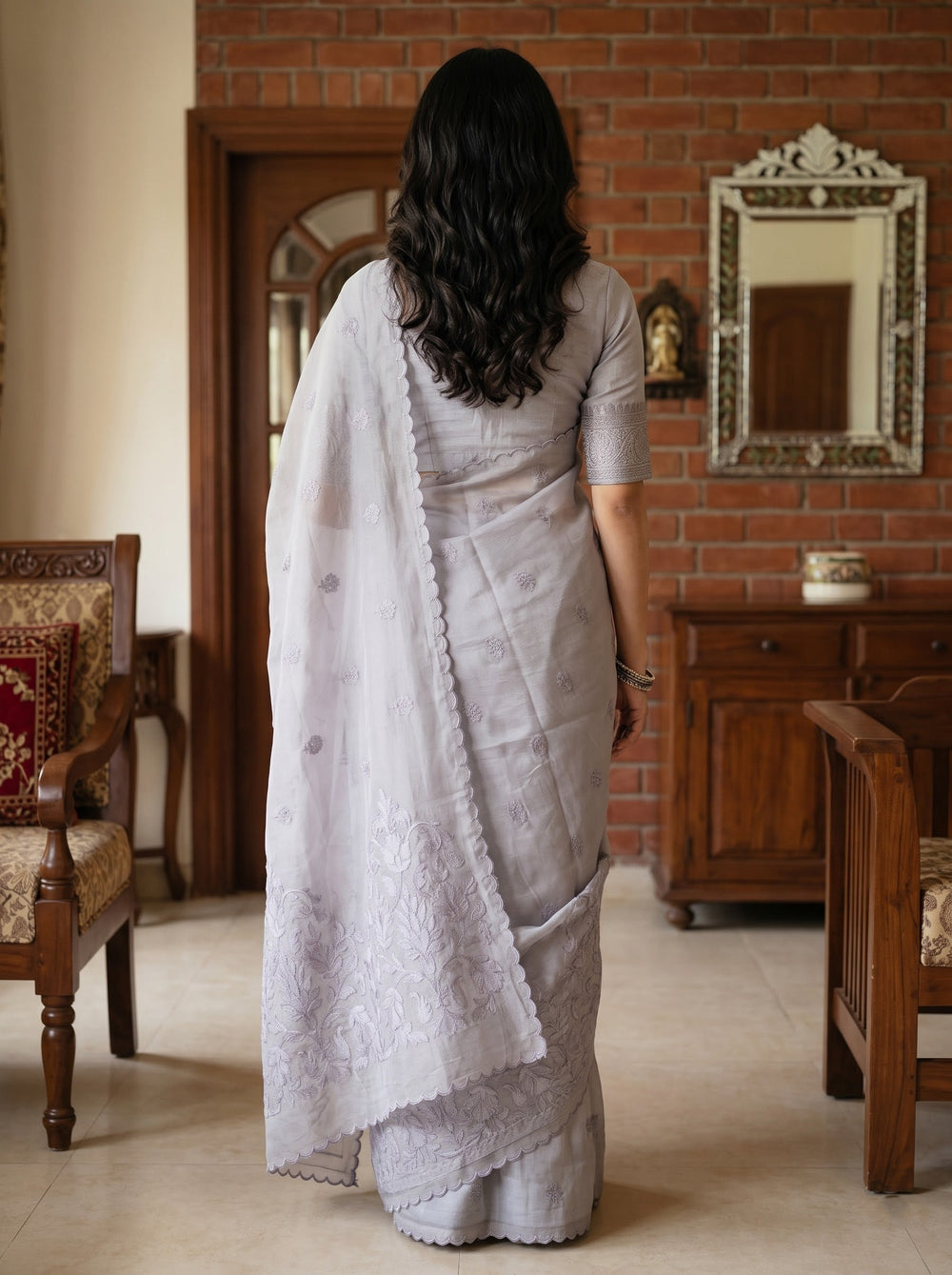 A woman wearing a light gray saree with a matching short-sleeve blouse, standing in a traditional indoor room with a brick wall, wooden furniture, and a mirror.