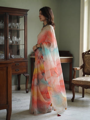 A woman wearing a flowing sheer saree with a watercolor-style print in coral, pink, orange, yellow, and light blue, standing in a vintage room with wooden furniture.