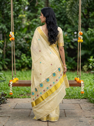 A woman wearing a cream-colored saree with gold borders and blue floral motifs, standing on a wooden swing in a lush green garden.