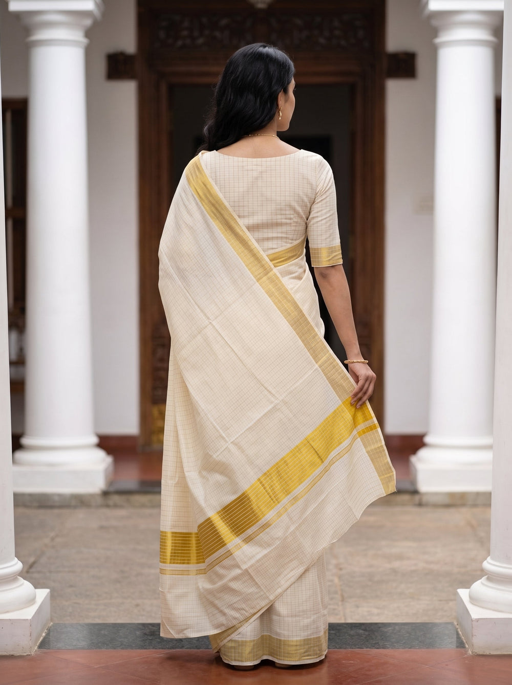 A woman wearing a cream-colored cotton saree with a mustard yellow border and matching blouse, standing in a courtyard with white pillars.