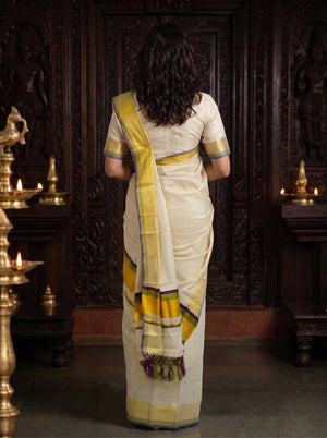 A woman viewed from behind wearing a cream-colored saree with yellow and purple border accents, standing in front of a dark carved wooden door with brass lamps on either side.