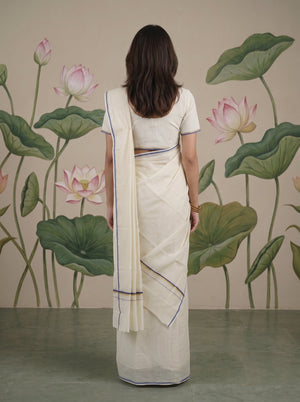 A woman viewed from behind wearing a cream-colored cotton saree with thin blue border stripes, paired with a matching cream blouse. She stands in front of a wall decorated with a large-scale green lotus leaf and pink lotus flower mural.