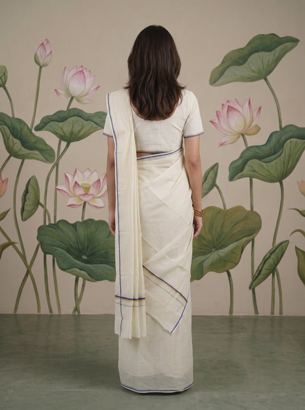 A woman viewed from behind wearing a cream-colored cotton saree with thin blue border stripes, paired with a matching cream blouse. She stands in front of a wall decorated with a large-scale green lotus leaf and pink lotus flower mural.