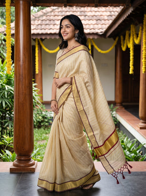 A woman modeling a beige and gold striped cotton saree with red border accents, standing in a traditional courtyard with wooden pillars and yellow garlands.