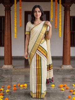 A woman modeling a cream-colored cotton saree with yellow, green, and red stripes, paired with a matching short-sleeve blouse, standing in a traditional courtyard with wooden pillars and hanging marigold garlands.