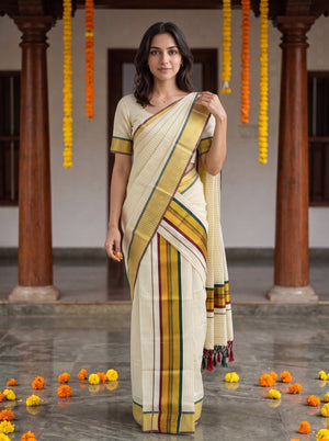 A woman modeling a cream-colored cotton saree with yellow, green, and red stripes, paired with a matching short-sleeve blouse, standing in a traditional courtyard with wooden pillars and hanging marigold garlands.