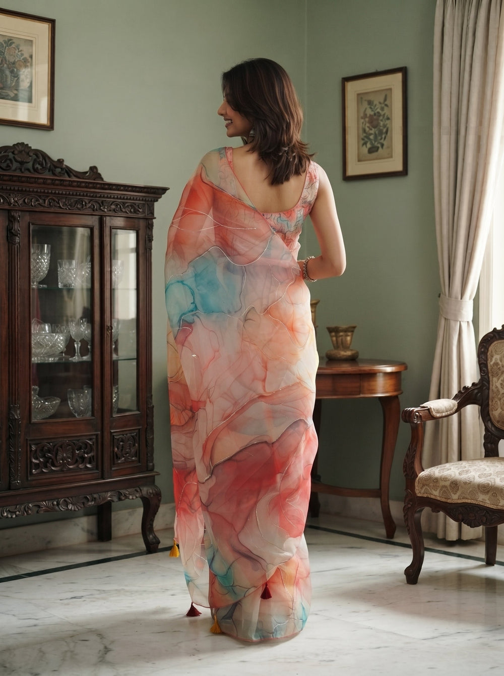 A woman wearing a semi-sheer peach and pink printed saree with yellow, orange, and blue watercolor-style floral patterns, standing in a vintage room with wooden furniture.