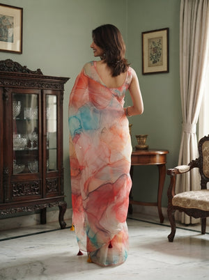 A woman stands in a vintage room wearing a sheer, semi-transparent saree with a watercolor floral pattern in pink, orange, and blue. The room features a dark wood cabinet, a small wooden table, and a chair against a sage green wall.