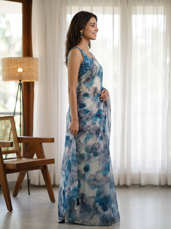 A woman wearing a sleeveless blue and white abstract-print maxi dress, standing in profile in a bright room with sheer white curtains, a wooden chair, and a floor lamp.