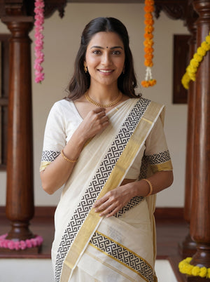 A woman wearing a traditional Indian saree with a cream body, black geometric border pattern, and gold trim, standing in a decorated indoor setting with wooden pillars and flower garlands.