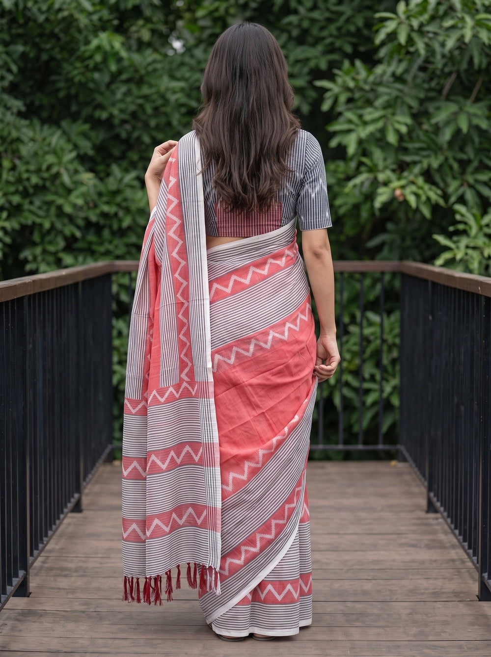 A woman wearing a coral pink and gray patterned saree with a matching gray printed blouse, standing on a wooden deck with green foliage in the background.