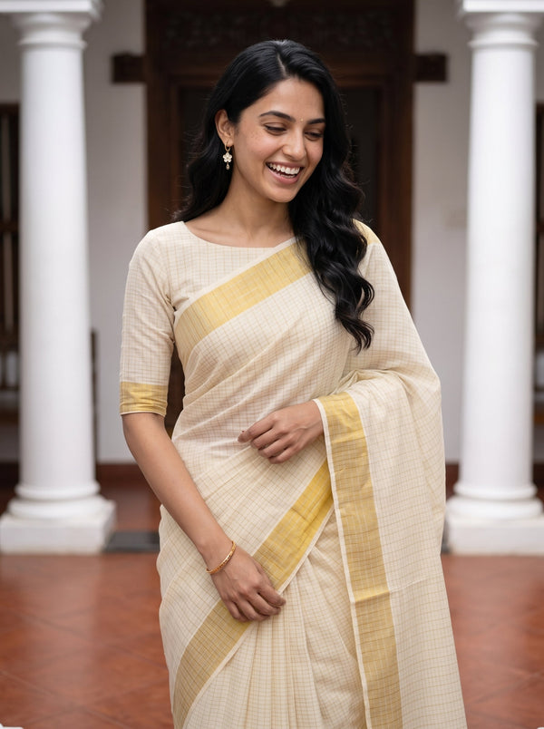 A woman wearing a cream-colored saree with a yellow border and matching yellow sleeve cuff, standing in front of white pillars.