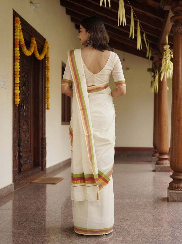 A woman wearing a white saree with green and orange stripes, viewed from behind as she stands in a traditional corridor with wooden pillars and garlands.