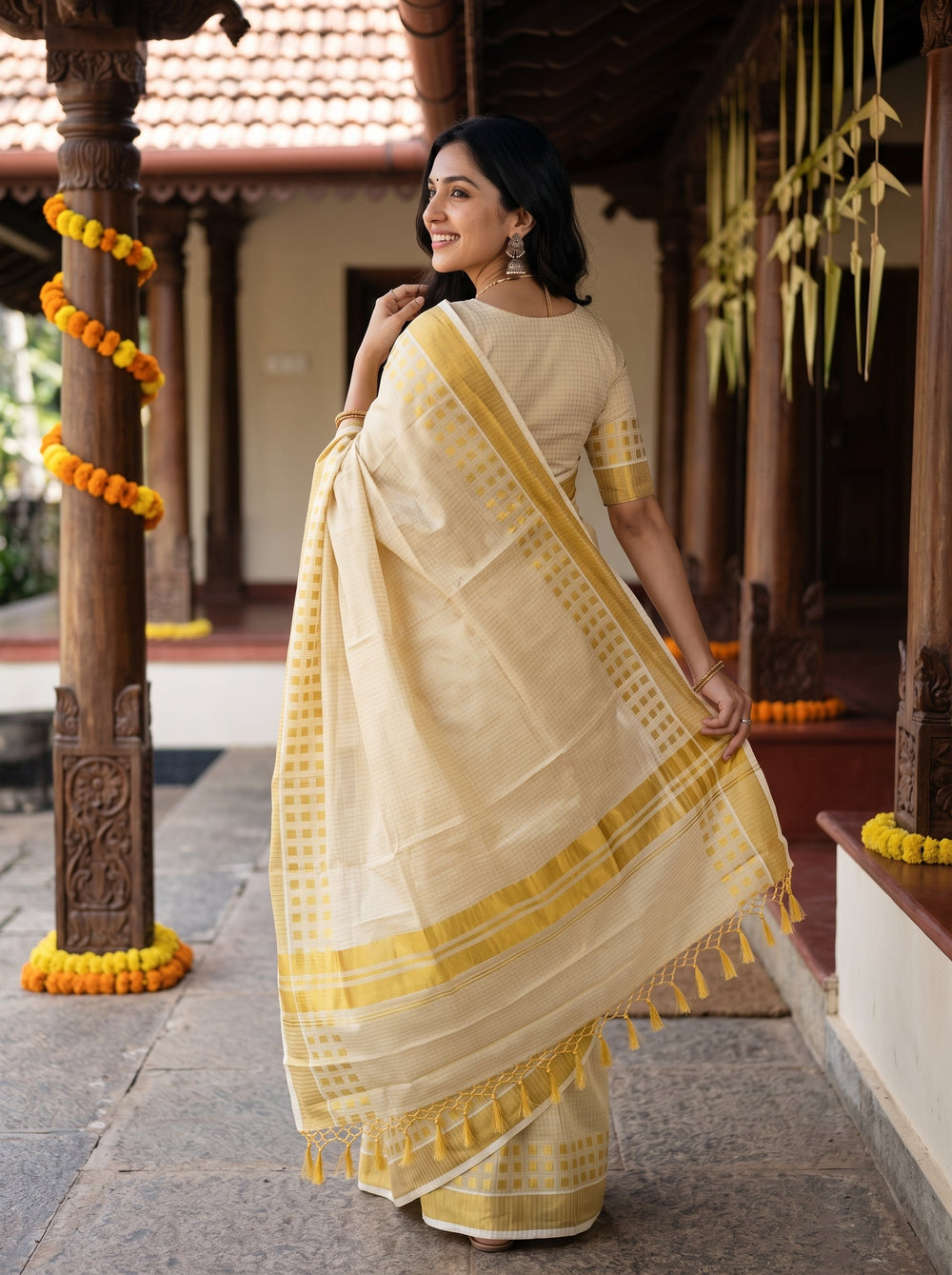 A woman wearing a light beige and yellow saree stands on steps in front of a decorated doorway with wooden pillars and marigold garlands.