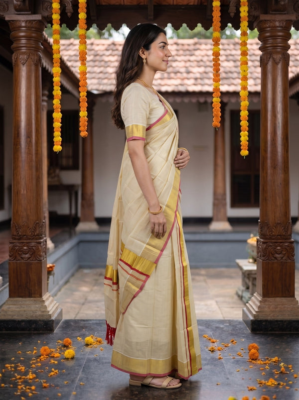 A woman wearing a cream-colored cotton saree with yellow and red border details, standing in profile in a traditional courtyard with wooden pillars and hanging marigold garlands.