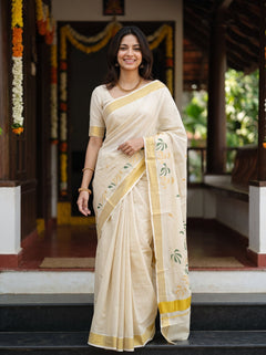 A woman wearing a cream-colored cotton saree with yellow borders and green floral motifs, standing in front of a traditional wooden doorway decorated with garlands.