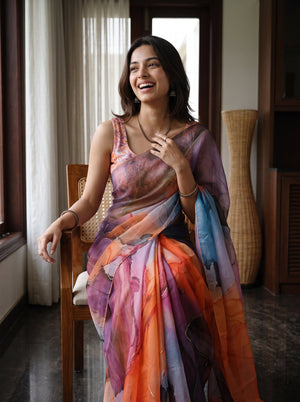 A woman wearing a colorful, semi-sheer sari with watercolor-like patterns in purple, orange, pink, and blue, sitting on a wooden chair in a home interior.