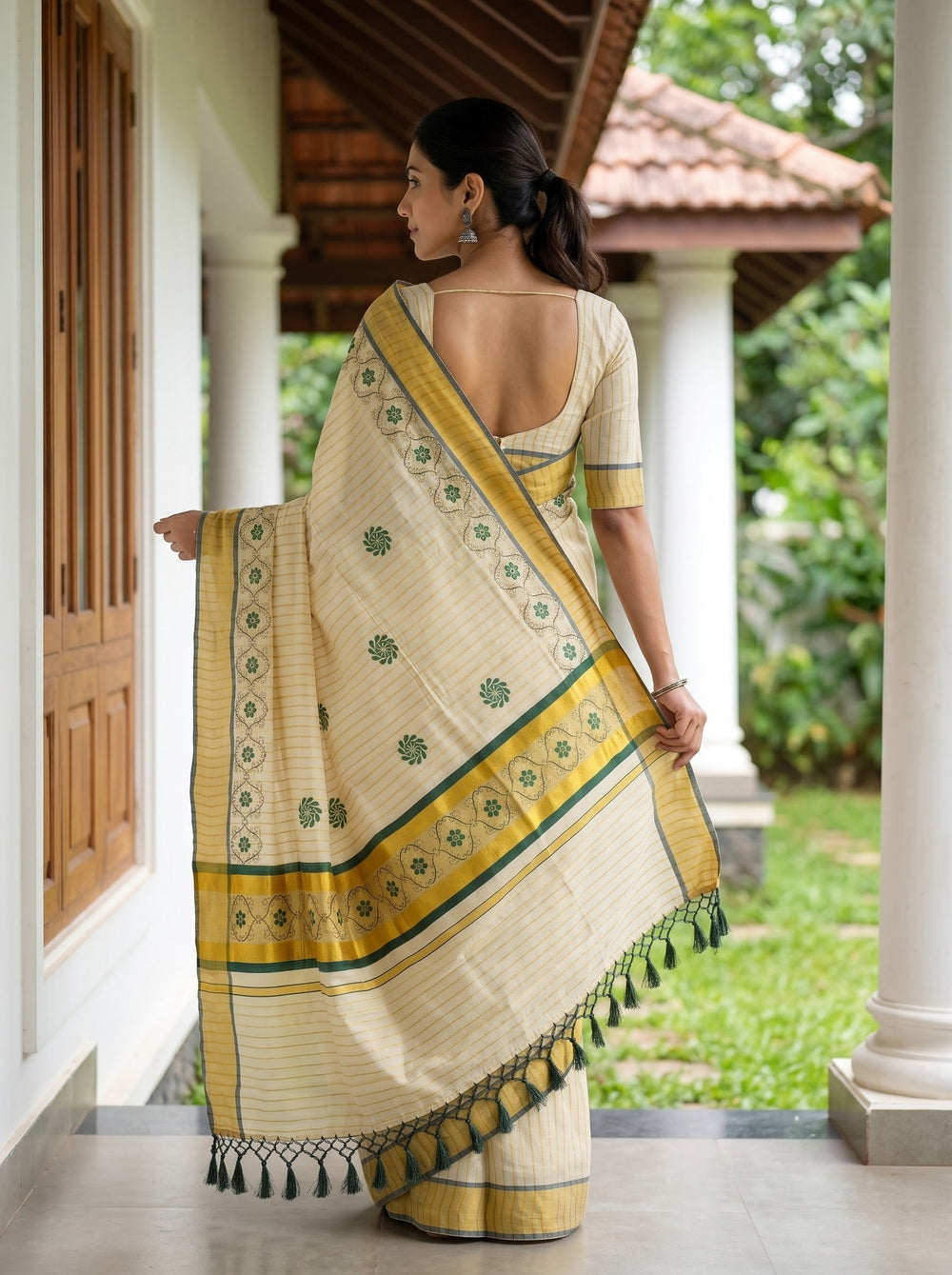 A woman wearing a cream-colored saree with yellow and green border details, standing on a porch with white pillars and a garden in the background.