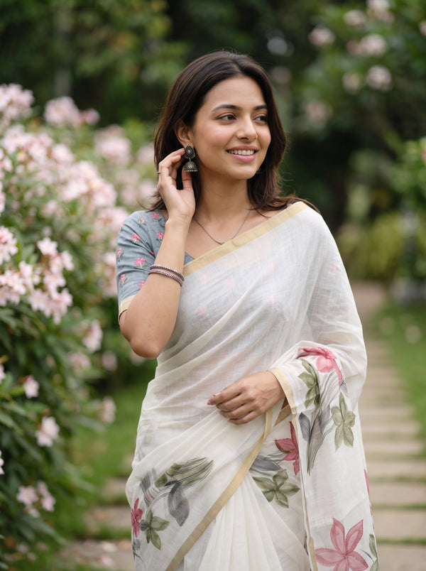 A woman wearing a white floral saree with a blue patterned blouse stands in a garden pathway, holding a phone to her ear.