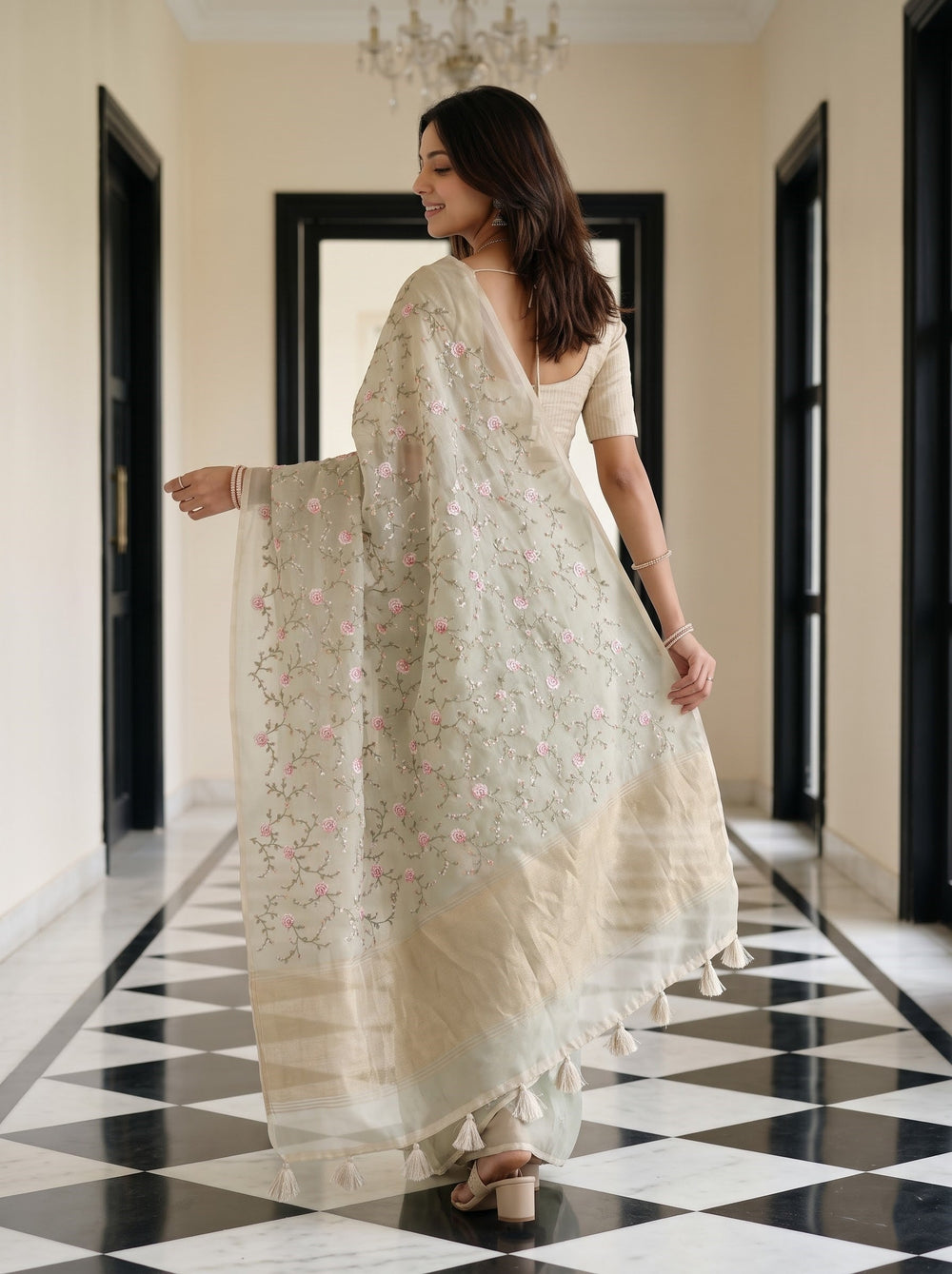 A woman wearing a light sage green saree with pink floral embroidery and a matching embroidered dupatta, standing in a hallway with a black and white checkered floor.