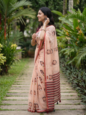 A woman wearing a peach-colored printed saree with red and dark stripes on the border, standing on a stone path surrounded by lush green tropical plants.