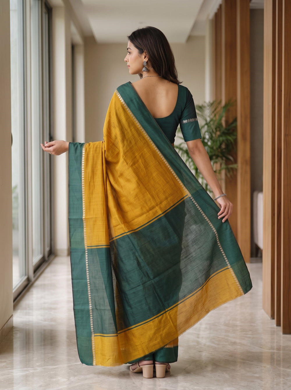 A woman wearing a mustard yellow and dark green saree with gold zari detailing, standing in a modern hallway with large windows and wooden slat walls.