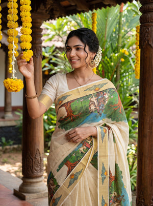 A woman wearing a cream-colored saree with a colorful traditional print stands under a wooden pavilion, holding yellow flower garlands.