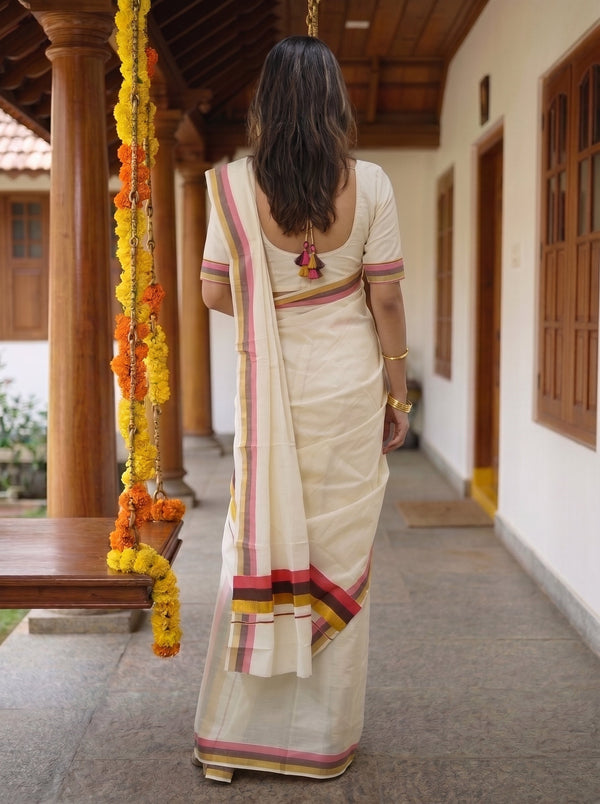 A woman viewed from behind wearing a cream-colored cotton saree with pink, yellow, and orange border stripes, standing in a traditional courtyard with wooden pillars and hanging marigold garlands.