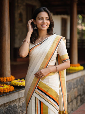 A woman wearing a cream-colored cotton saree with orange, yellow, and black striped borders, standing in front of a traditional wooden porch with flower garlands.