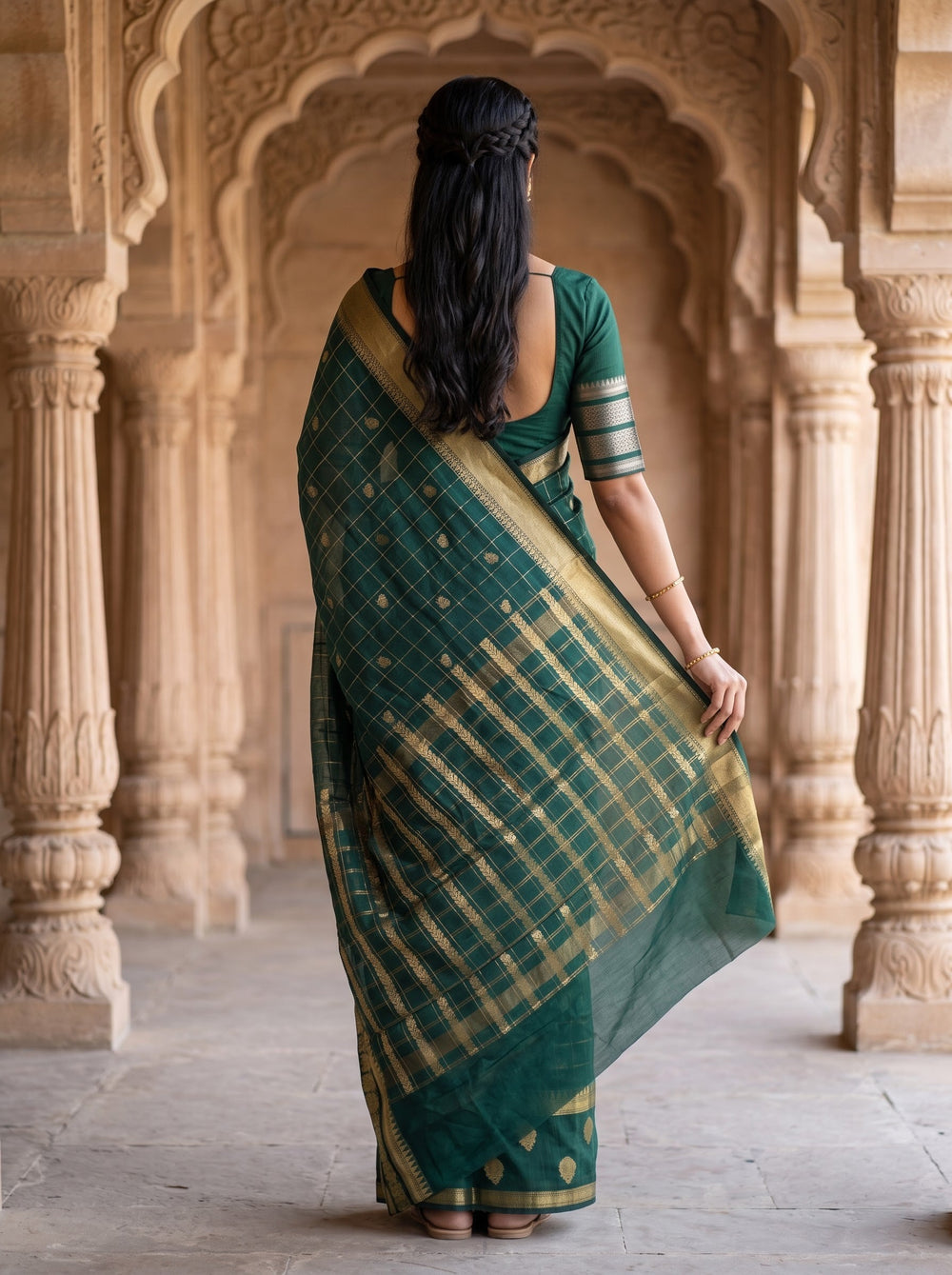 A woman wearing a dark green silk saree with gold zari patterns and a matching green blouse, standing in front of an ornate beige stone archway.