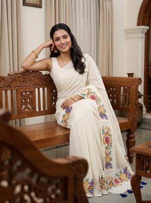 A woman wearing a white saree with colorful floral embroidery sits on a carved wooden bench in an indoor setting with beige curtains.