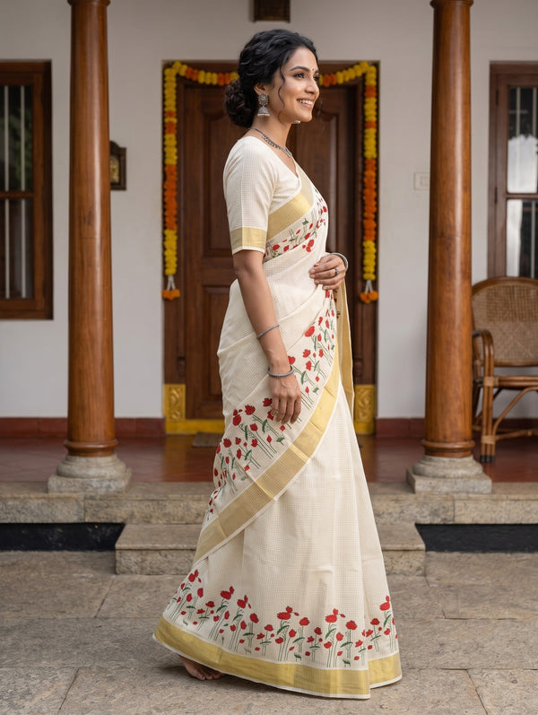 A woman wearing a cream-colored cotton saree with red floral embroidery and gold borders, standing in a traditional courtyard with wooden pillars.