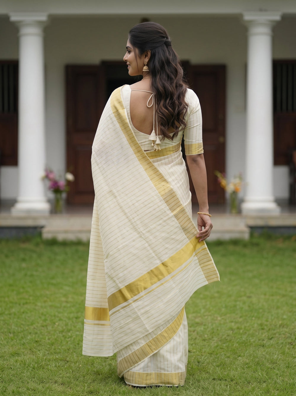 A woman wearing a cream-colored saree with gold stripes and a gold border, standing on a green lawn in front of a house with white columns.