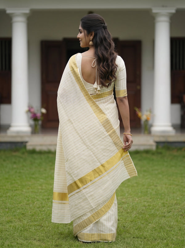 A woman wearing a cream-colored saree with gold stripes and a matching cream blouse, standing outdoors on a green lawn in front of a house with white columns.