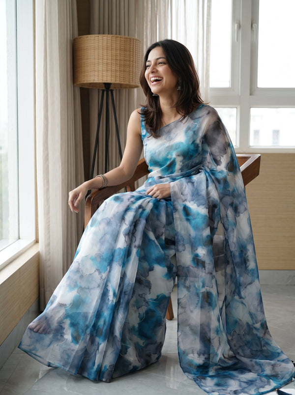 A woman wearing a sleeveless, flowing maxi dress with a blue, gray, and white watercolor-style print, sitting on a wooden chair in a bright room with large windows and a floor lamp.