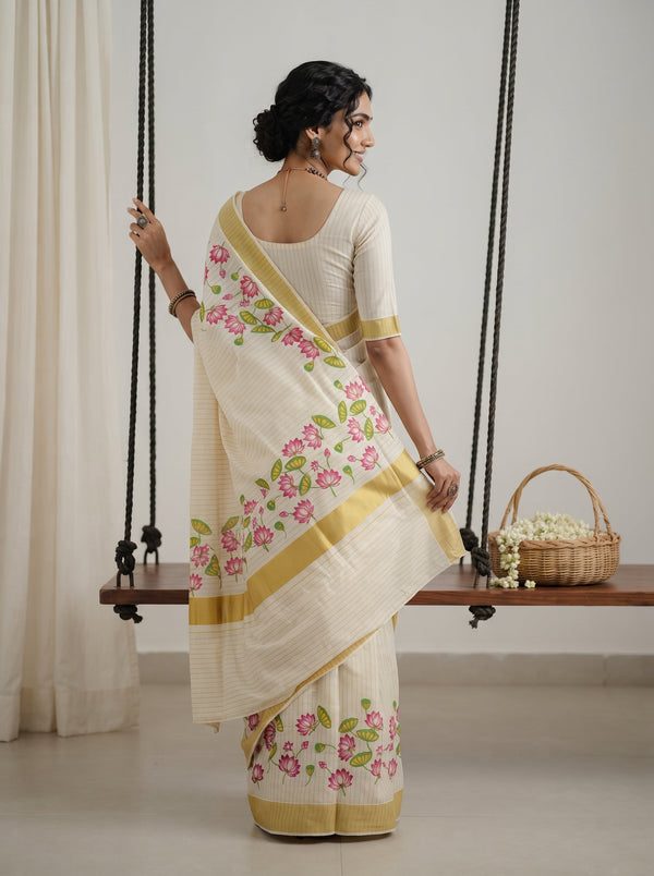 A woman wearing a cream-colored saree with pink floral prints and gold borders, standing in front of a wooden swing in a studio setting.