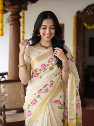 A woman wearing a cream-colored cotton saree with pink lotus floral prints and a gold border, standing in a traditional indoor setting with wooden pillars.