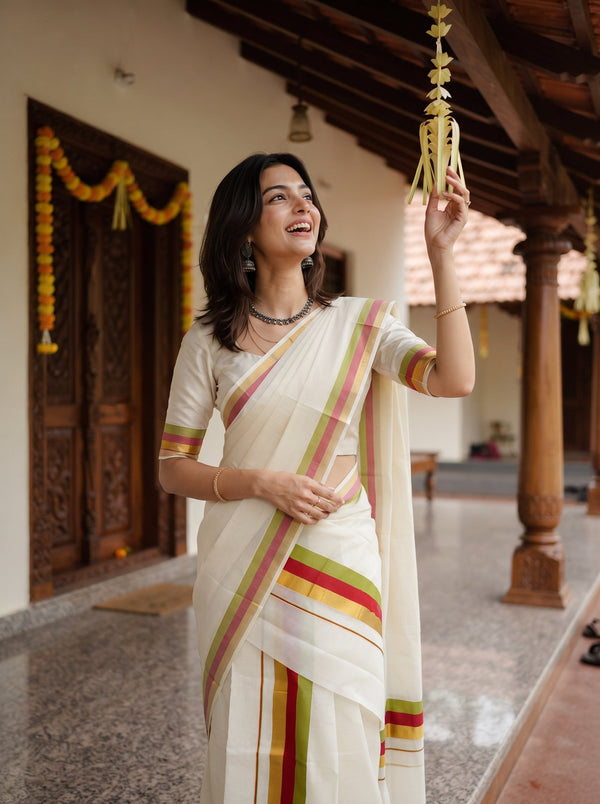 A woman wearing a white saree with green, yellow, and red stripes stands in a traditional courtyard, holding a hanging decorative garland.