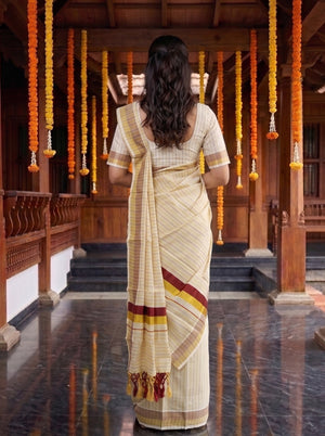 A woman viewed from behind wearing a cream-colored striped saree with maroon and yellow border accents, standing in a traditional wooden corridor decorated with orange and yellow marigold garlands.
