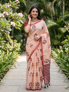 A woman stands on a stone path surrounded by lush green plants, wearing a peach-colored printed saree with a red border and a matching red blouse.
