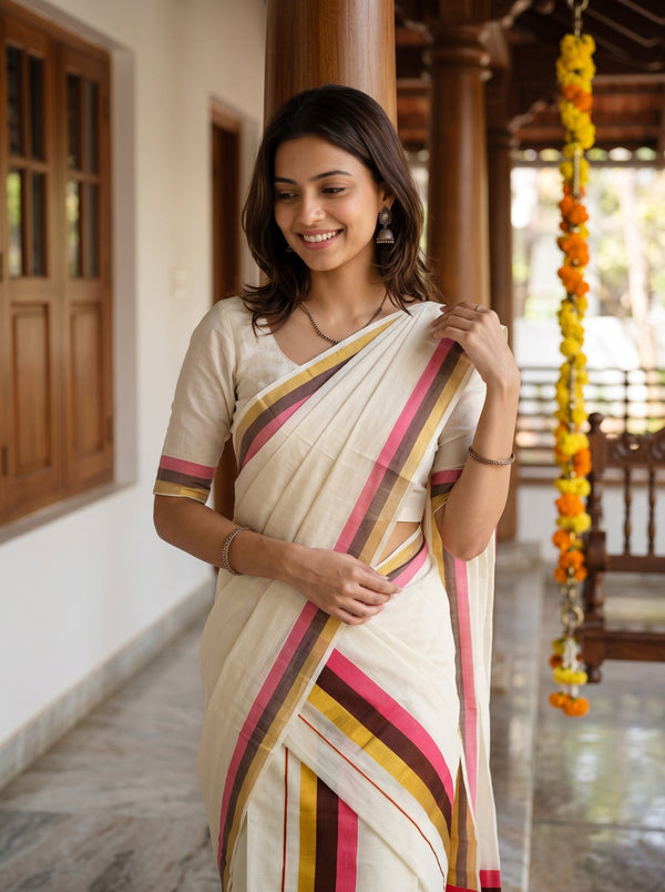 A woman wearing a cream-colored cotton saree with pink, mustard yellow, and brown striped borders, standing in a traditional house corridor.
