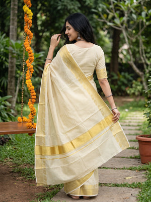 A woman wearing a cream-colored saree with yellow-gold borders and a textured pattern, standing outdoors in a garden setting.
