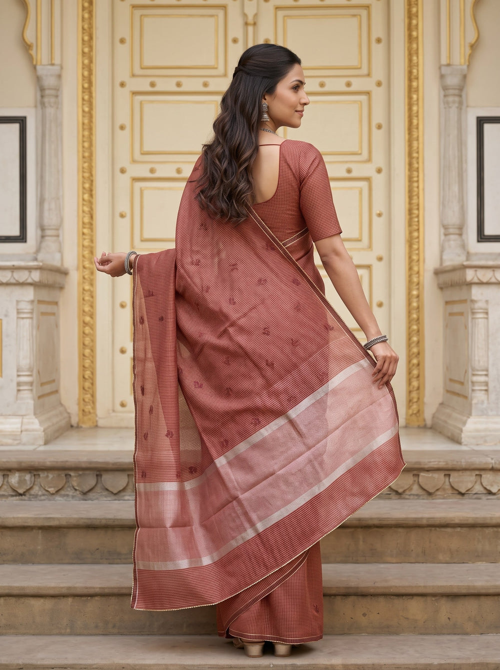 A woman wearing a dusty rose terracotta cotton saree with a matching printed blouse, standing on stone steps in front of an ornate cream-colored building door.