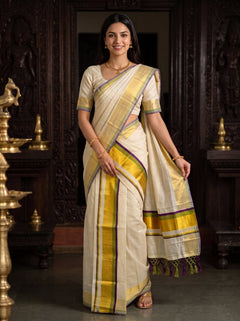 A woman modeling a cream-colored silk saree with yellow, purple, and green borders, standing in front of a dark carved wooden door with brass lamps on either side.