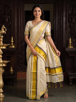 A woman modeling a cream-colored silk saree with yellow, purple, and green borders, standing in front of a dark carved wooden door with brass lamps on either side.