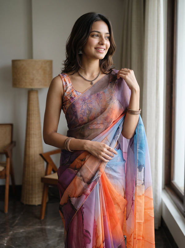 A woman wearing a colorful printed saree with a matching sleeveless blouse, standing indoors near a window. The saree has an abstract watercolor-like pattern in shades of purple, blue, orange, and pink.