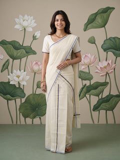 A woman modeling a cream-colored cotton saree with thin blue and green border stripes, paired with a matching cream blouse, standing in front of a painted lotus wall mural.