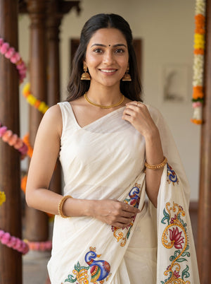 A woman wearing a white silk saree with colorful embroidered motifs, standing in a decorated indoor setting with garlands.