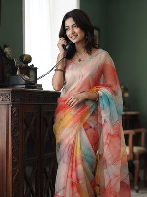 A woman wearing a lightweight multicolor printed saree stands indoors near a dark wooden cabinet, holding a black rotary telephone. The saree has an abstract watercolor-like pattern in soft pink, orange, yellow, blue, and white tones.